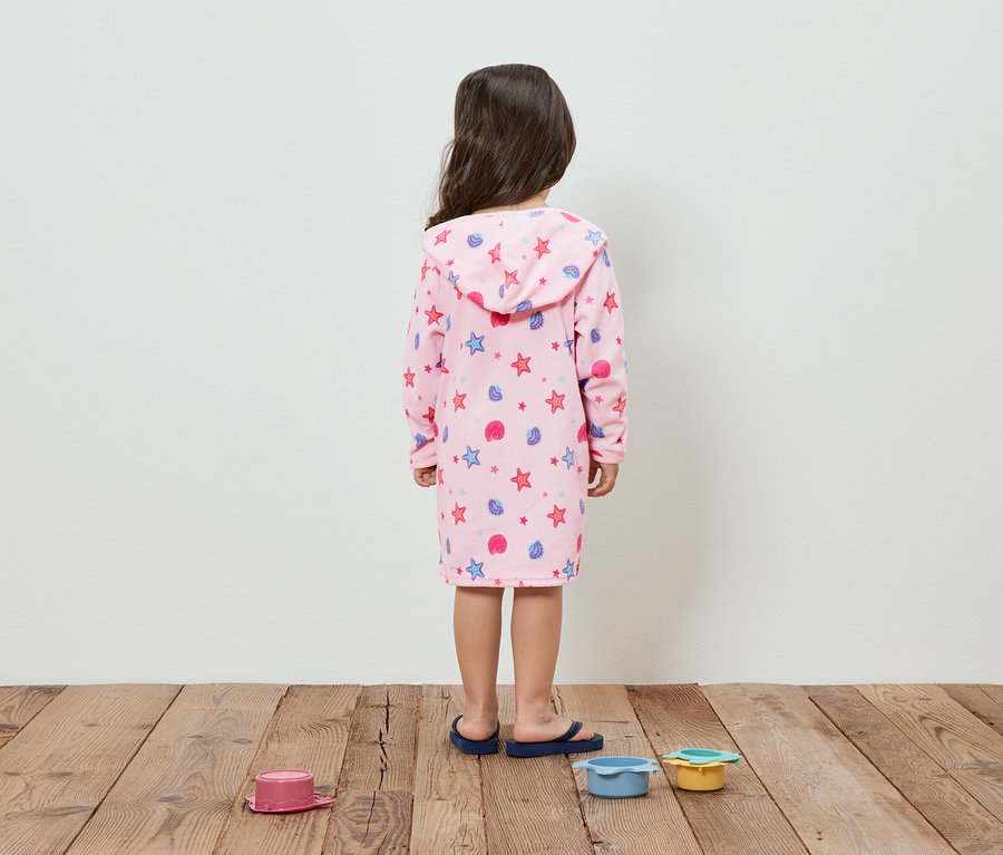 Un enfant vu de dos, portant un peignoir rose avec des étoiles et des coquillages, se tient sur un plancher en bois à côté de jouets.