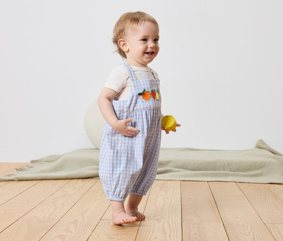 Un jeune enfant se tient pieds nus sur un plancher en bois et tient un bloc de jeu jaune dans sa main. L'enfant porte une salopette à carreaux bleu et blanc avec des motifs de fruits.
