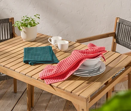 Sur une table en bois, deux tasses blanches, un torchon gaufré vert et un rose, des assiettes et un bol.