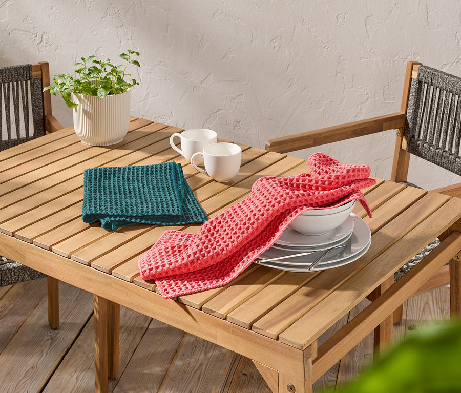 Sur une table en bois, deux tasses blanches, un torchon gaufré vert et un rose, des assiettes et un bol.
