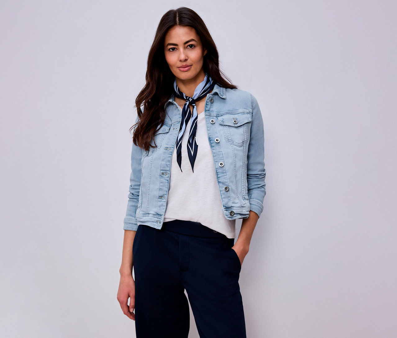 Femme portant une veste en jean bleu clair, un t-shirt blanc, un pantalon bleu marine et un foulard bleu et blanc sur fond clair.