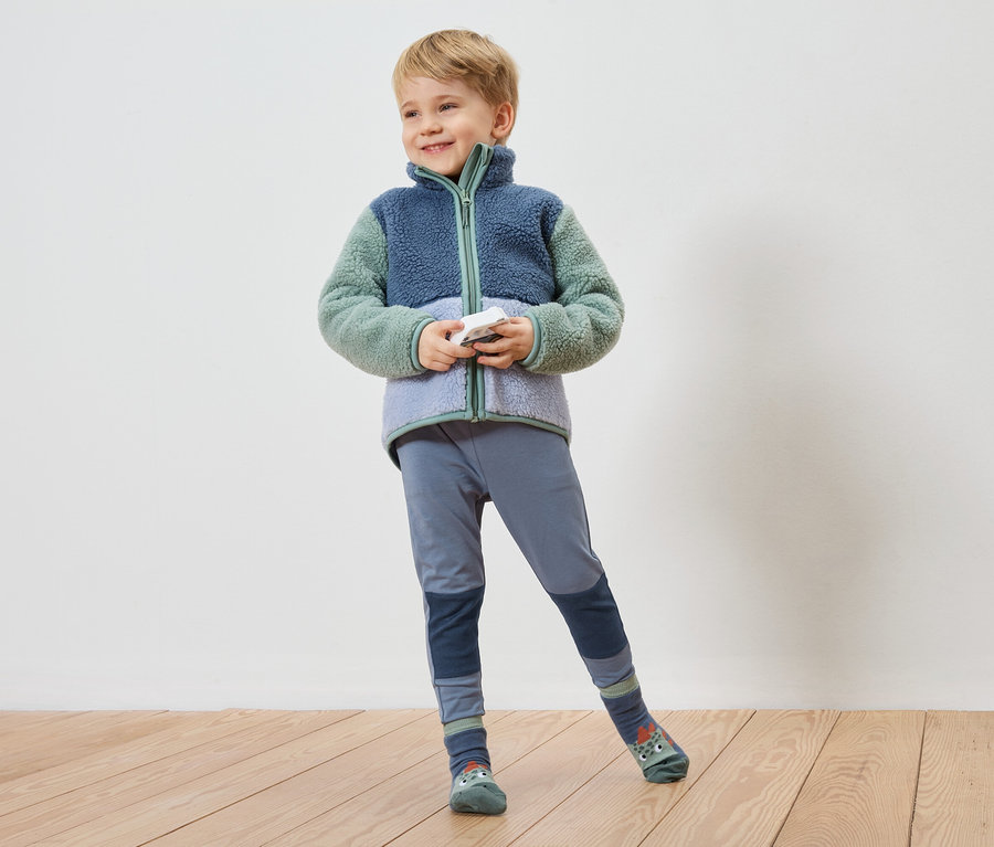 Un garçon souriant aux cheveux blonds courts pose devant un mur, vêtu d'une veste en polaire bleu-vert, d'un legging bleu et de chaussettes à motifs d'animaux.