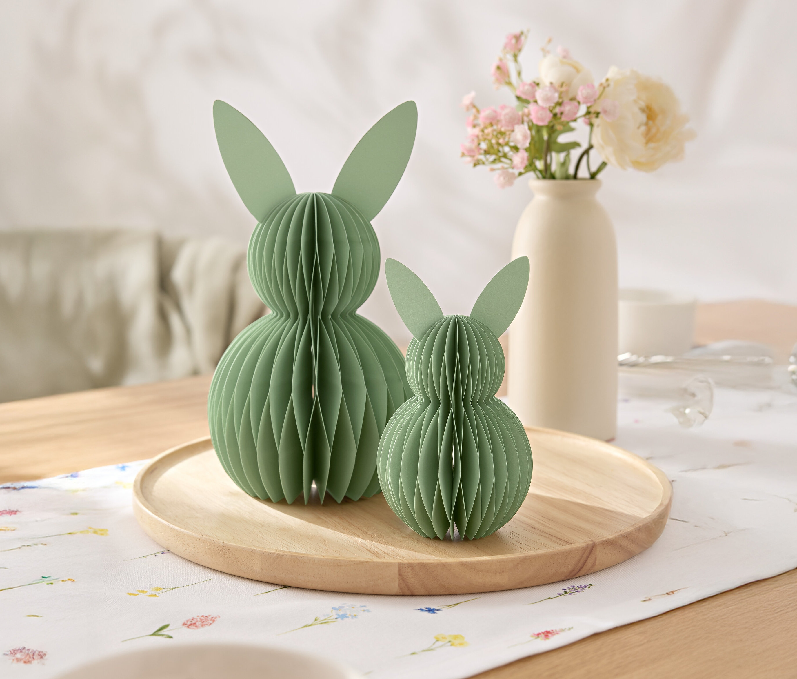Deux lapins en papier vert sont posés sur un plateau en bois, avec un vase de fleurs en arrière-plan.