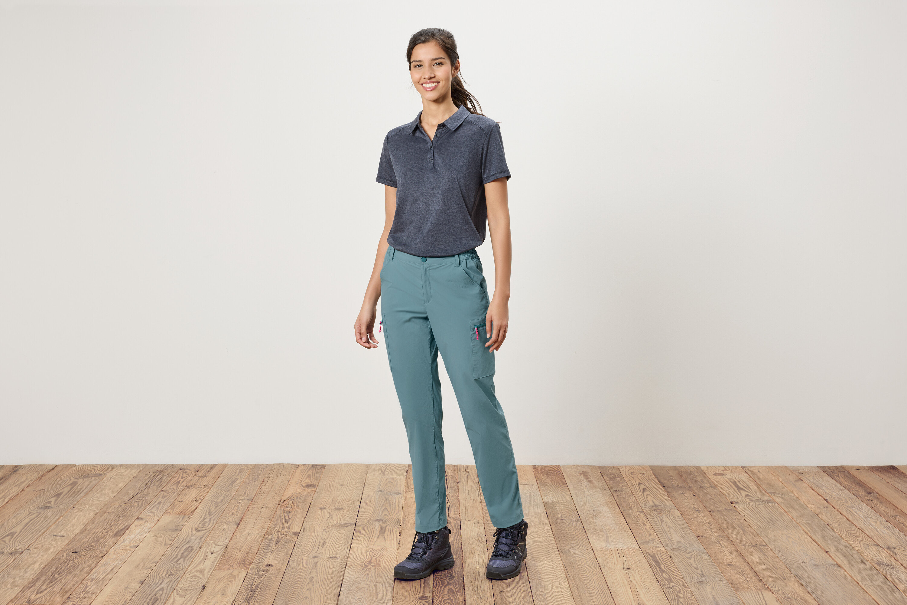 Une femme se tient devant un mur blanc, portant un polo bleu, un pantalon bleu et des chaussures de randonnée.