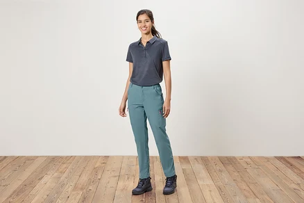 Une femme se tient devant un mur blanc, portant un polo bleu, un pantalon bleu et des chaussures de randonnée.