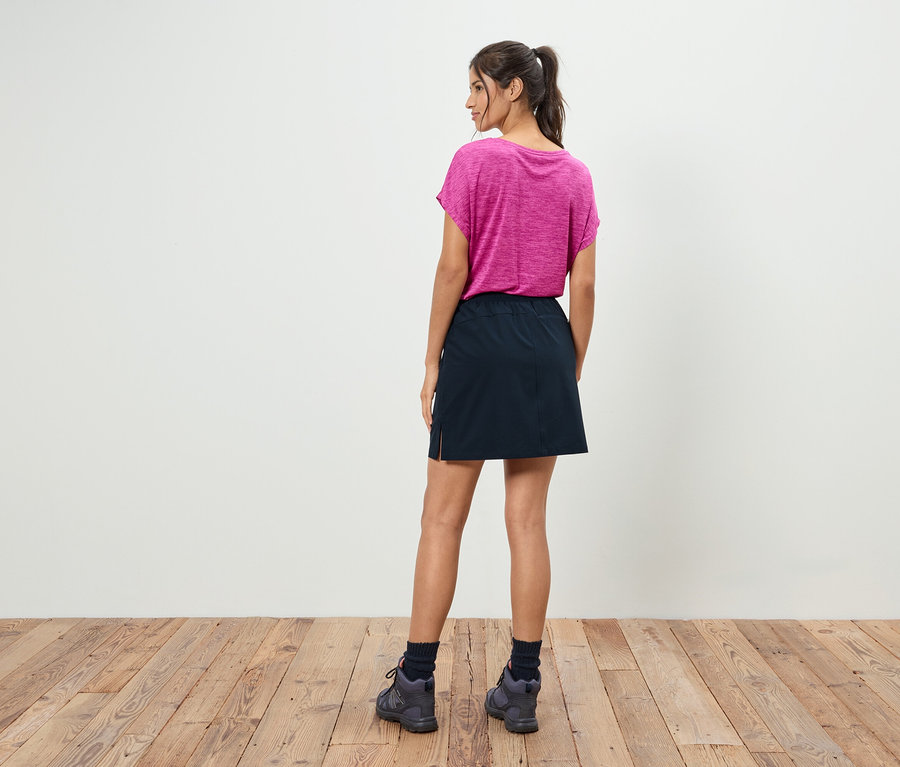 Une femme avec une queue de cheval se tient dos à l'appareil photo et porte un t-shirt rose, une jupe noire et des chaussures grises.