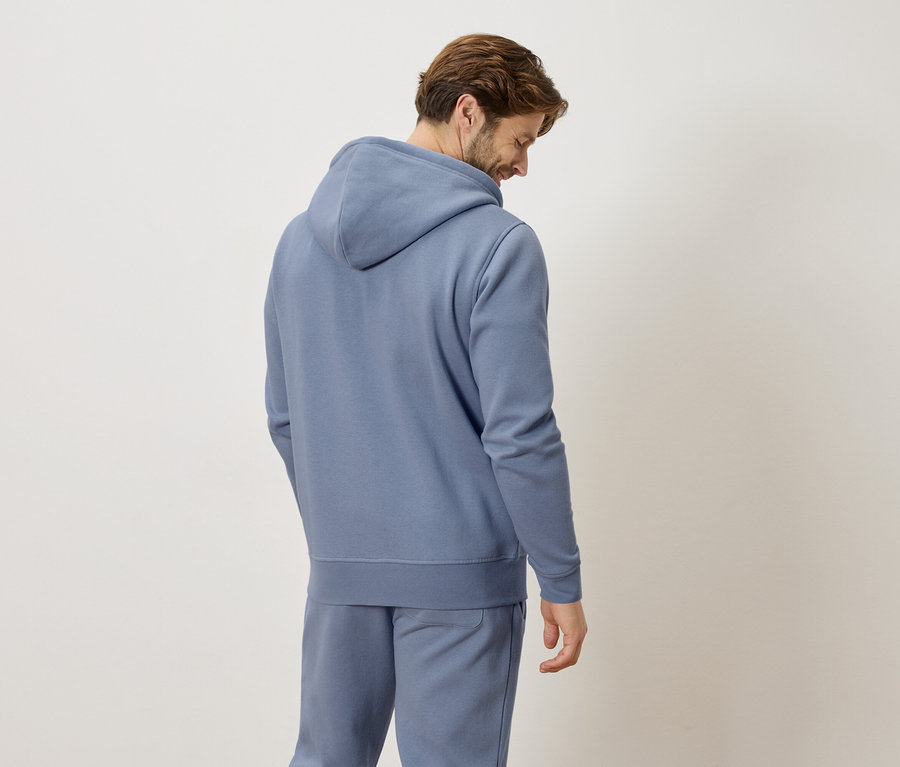 Homme vu de dos portant un sweat à capuche bleu et un pantalon de jogging sur un fond neutre.