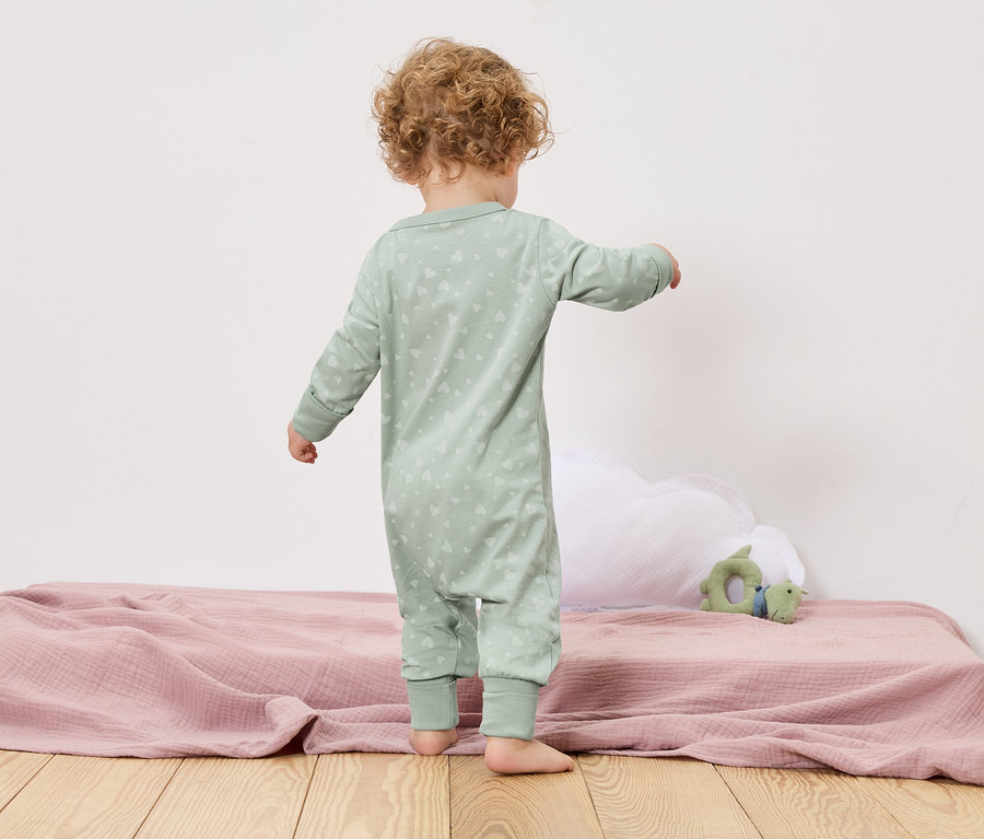 Un jeune enfant se tient dos à la caméra sur un lit avec une couverture rose et un jouet vert.