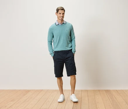 Un homme debout, les mains dans les poches, devant un mur, portant un pull vert menthe, un short cargo et des baskets blanches.