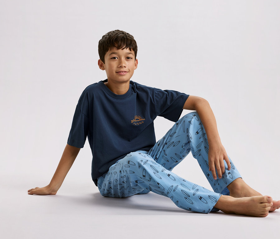 Un garçon est assis par terre, portant un t-shirt bleu marine avec un petit soleil et des vagues dessus et un pantalon de pyjama bleu clair avec un motif de planches de surf.
