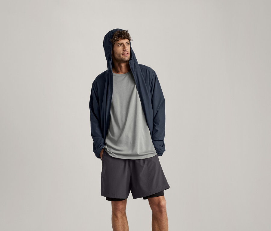 Un homme porte un t-shirt de sport gris clair chiné, une veste à capuche bleu marine et un short.