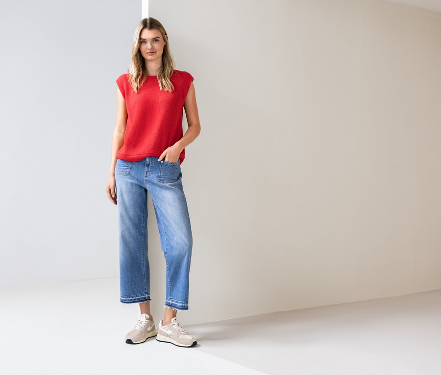 Femme debout dans une pièce lumineuse, portant un haut rouge, un jean bleu et des baskets.