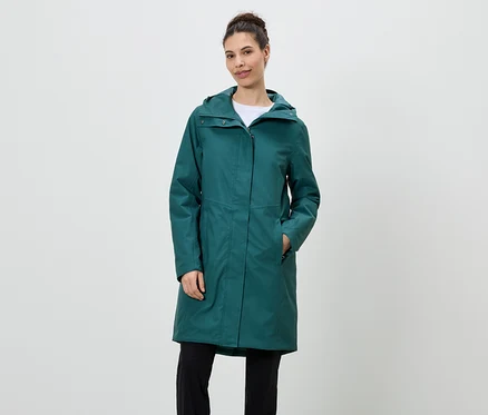 Femme debout portant un imperméable vert émeraude à capuche et un pantalon noir.