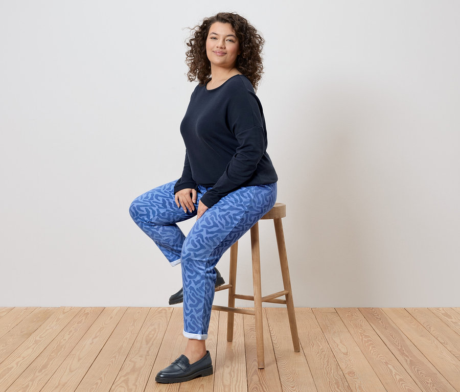 Une femme aux cheveux bouclés est assise sur un tabouret, portant un haut bleu marine, un pantalon à motifs bleus et des mocassins noirs.