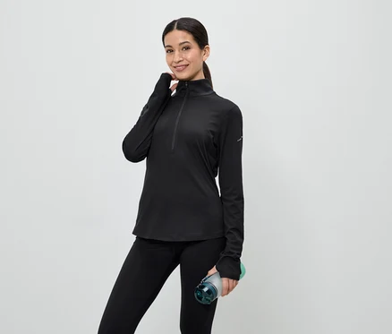 Une femme portant un haut de sport noir zippé et un legging tient une bouteille d'eau devant un fond clair.
