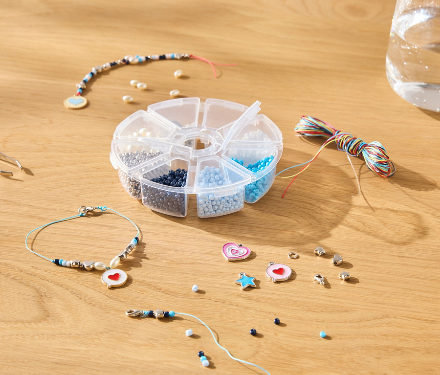 Sur une table en bois sont posés une pince à épiler, une boîte de perles, des fils colorés, une bouteille d'eau et des bijoux.