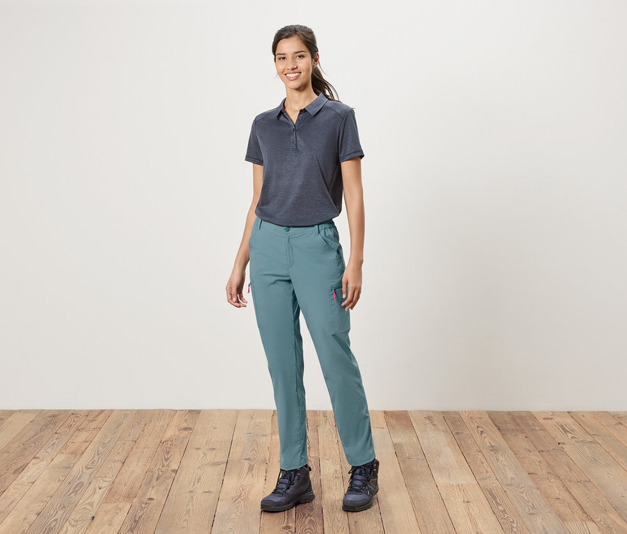 Une femme se tient devant un mur blanc, portant un polo bleu, un pantalon bleu et des chaussures de randonnée.