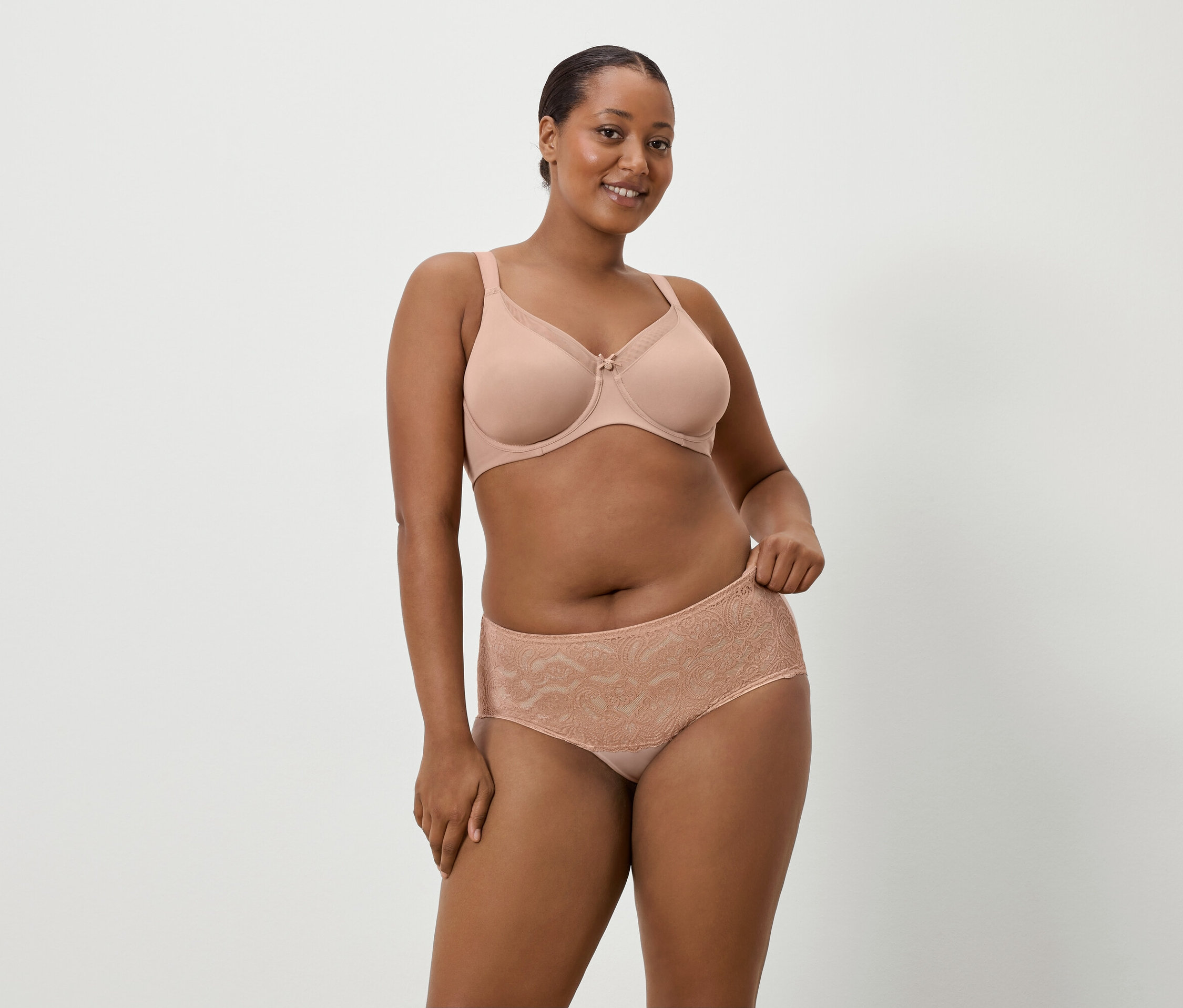 Une femme à la peau foncée porte un soutien-gorge couleur chair et une culotte en dentelle assortie sur un fond uni.