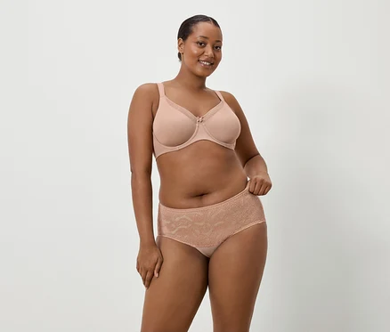 Une femme à la peau foncée porte un soutien-gorge couleur chair et une culotte en dentelle assortie sur un fond uni.