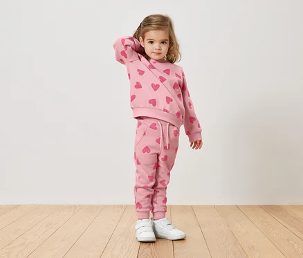 Une fillette blonde porte un pull rose et un pantalon avec un motif de cœur et des baskets blanches.