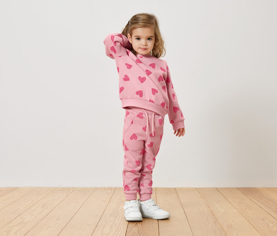 Une fillette blonde porte un pull rose et un pantalon avec un motif de cœur et des baskets blanches.