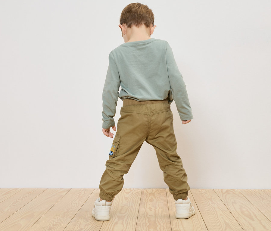 Un enfant se tient dos à la caméra, vêtu d'un t-shirt vert à manches longues, d'un pantalon kaki et de baskets blanches.