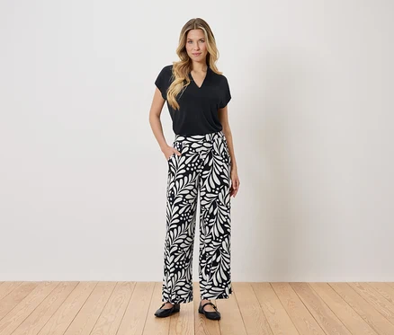 Une femme se tient debout, une main dans la poche, devant un mur blanc. Elle porte un chemisier noir et un pantalon noir et blanc.