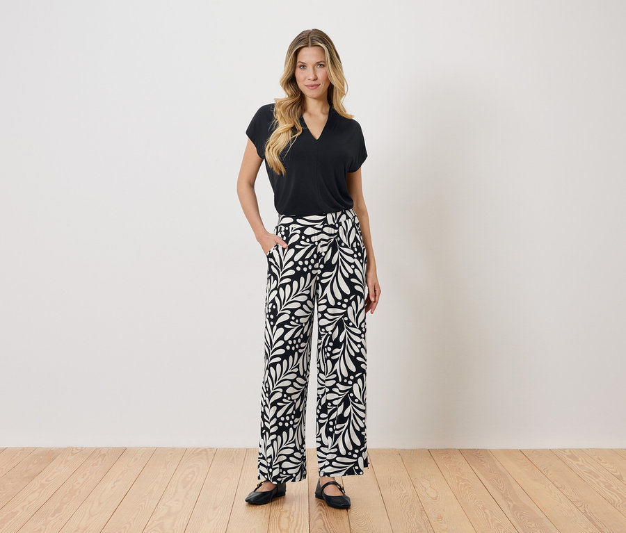 Une femme se tient debout, une main dans la poche, devant un mur blanc. Elle porte un chemisier noir et un pantalon noir et blanc.