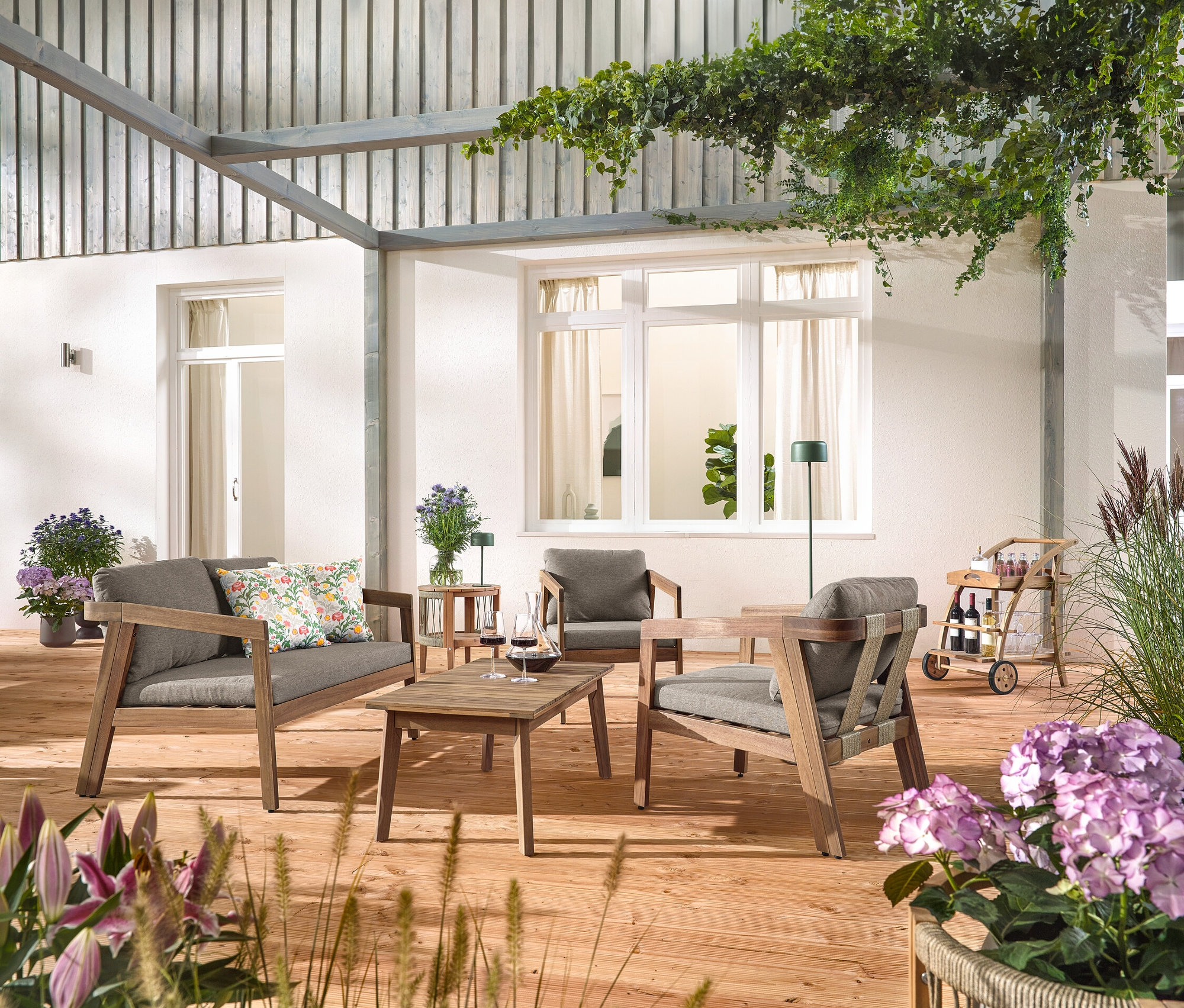 Sur une terrasse en bois se trouve un ensemble de salon de jardin avec tresse textile, 4 pièces avec 2 coussins d'extérieur à motifs floraux, un barwagen avec plateau amovible et une table d'appoint de jardin avec plateau amovible, Ø env. 50 cm. Une lampe de table à accumulateur solaire, vert jungle et une lampe de sol à accumulateur solaire, vert jungle sont également visibles.