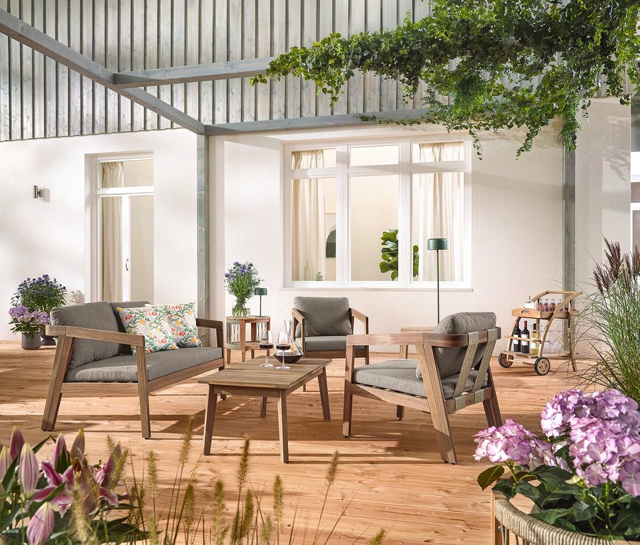 Sur une terrasse en bois se trouve un ensemble de salon de jardin avec tresse textile, 4 pièces avec 2 coussins d'extérieur à motifs floraux, un barwagen avec plateau amovible et une table d'appoint de jardin avec plateau amovible, Ø env. 50 cm. Une lampe de table à accumulateur solaire, vert jungle et une lampe de sol à accumulateur solaire, vert jungle sont également visibles.