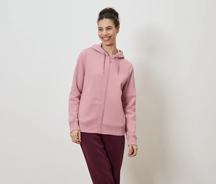 Une femme souriante portant les cheveux en chignon porte un sweat à capuche rose clair zippé et un pantalon bordeaux sur fond blanc.