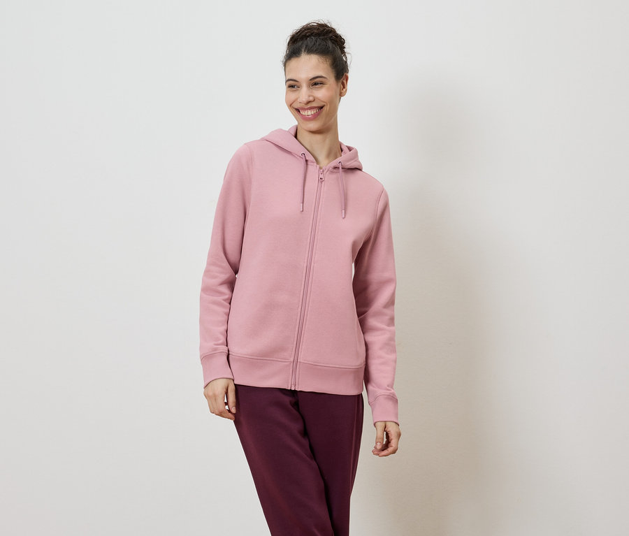 Une femme souriante portant les cheveux en chignon porte un sweat à capuche rose clair zippé et un pantalon bordeaux sur fond blanc.