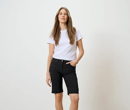 Une femme aux longs cheveux bruns, portant un t-shirt blanc et un short noir, pose avec une main dans la poche devant un fond blanc.