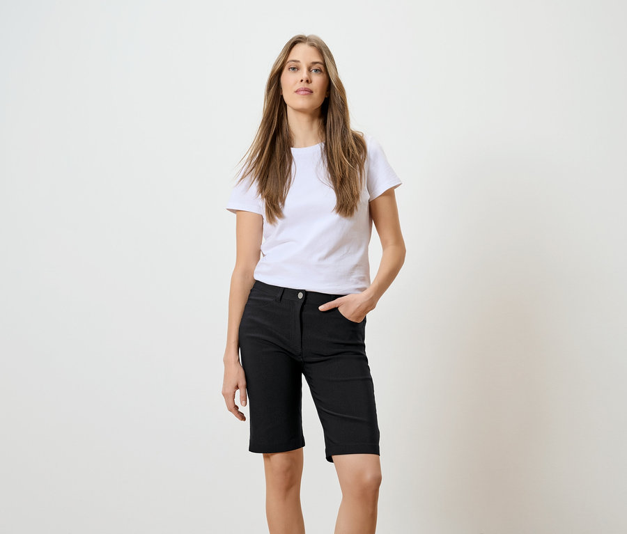 Une femme aux longs cheveux bruns, portant un t-shirt blanc et un short noir, pose avec une main dans la poche devant un fond blanc.