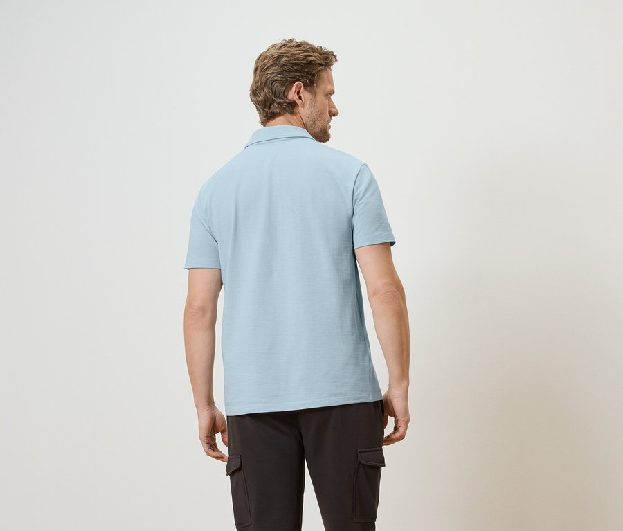 Homme vu de dos portant un polo bleu clair et un pantalon cargo foncé sur un fond clair.