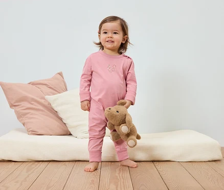 Un bambin souriant en pyjama rose tient un ours en peluche devant un lit avec des coussins.