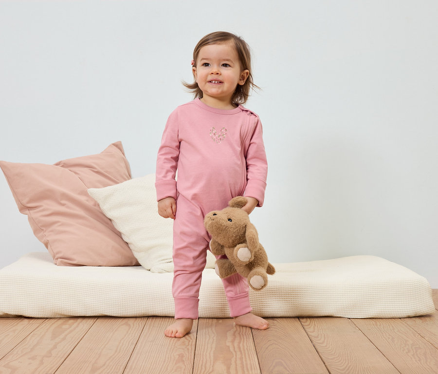 Un bambin souriant en pyjama rose tient un ours en peluche devant un lit avec des coussins.