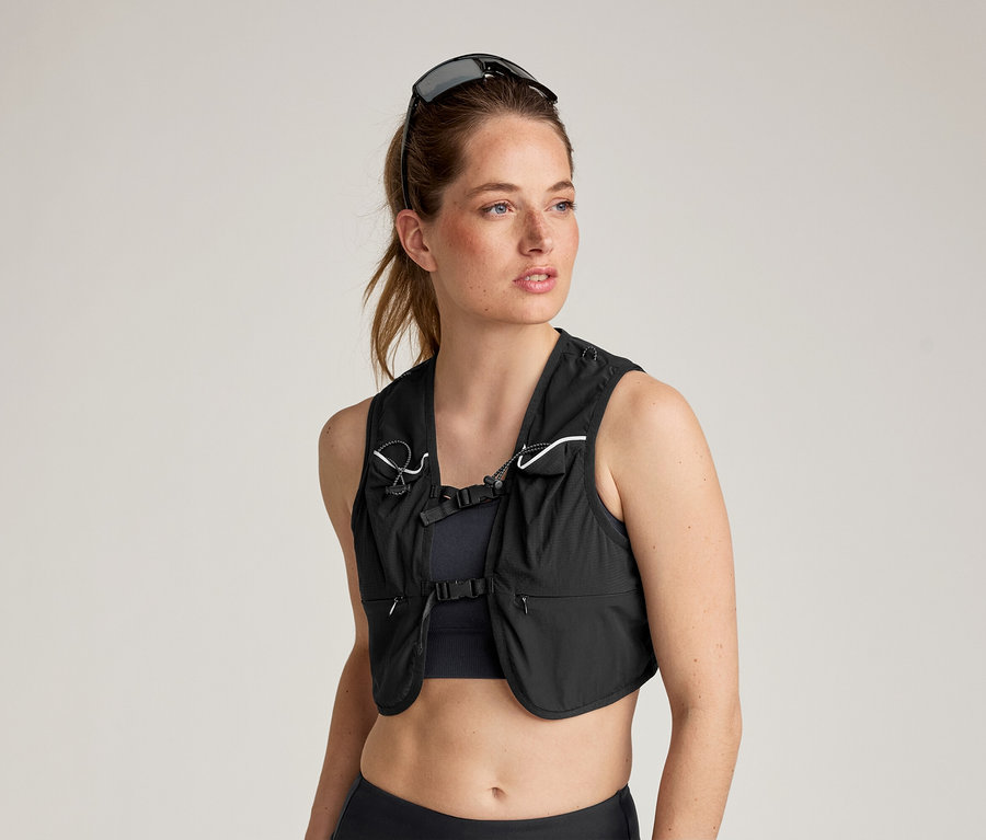 Une femme portant des lunettes de soleil sur la tête porte un gilet de course noir sur un soutien-gorge de sport et un legging.