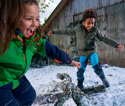 Deux enfants jouent dans la neige. Une fille en veste verte rit et tend la main. Un garçon en veste kaki et pantalon de neige bleu est en équilibre sur une bûche enneigée. Un bâtiment en bois est en arrière-plan.