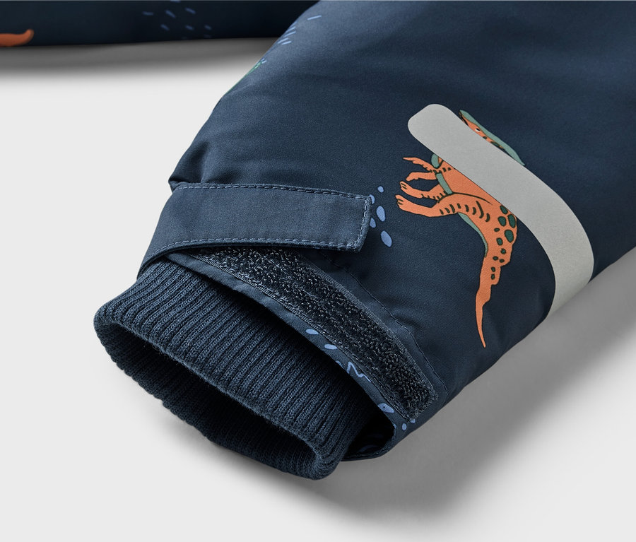 Gros plan sur la manche d'une veste bleu marine à motif de dinosaures, avec poignet côtelé et patte velcro.