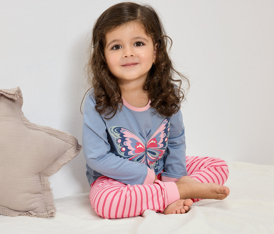 Une jeune fille aux cheveux bouclés est assise sur un lit et porte un pyjama bleu avec un motif de papillon et un pantalon rayé rose.