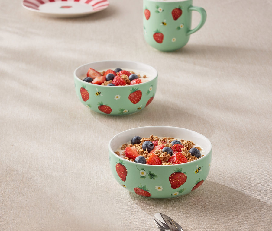 Sur une table, deux bols de granola et de baies, une tasse et une assiette, tous décorés de motifs de fraises et d'abeilles.