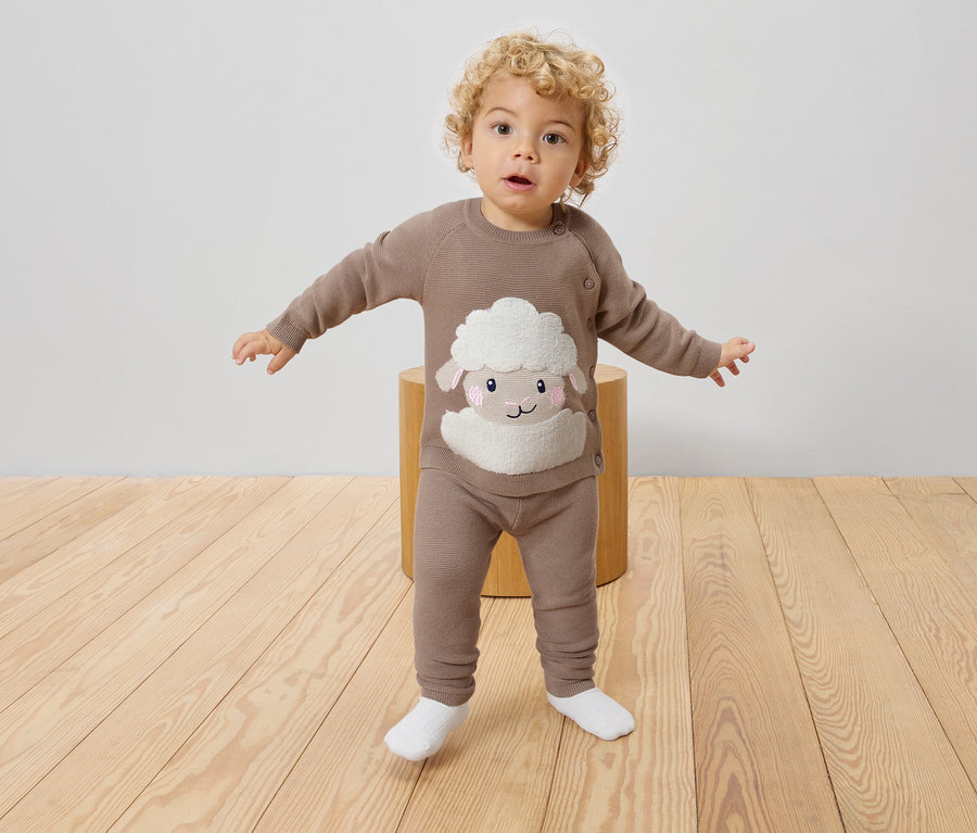 Un jeune enfant aux cheveux bouclés se tient sur un tabouret, portant un pull en tricot marron et un legging avec un mouton dessus.