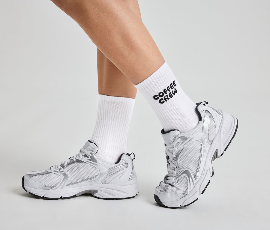 Jambes d'une personne portant des chaussettes blanches avec l'inscription "Coffee Crew" et des baskets blanches et argentées.