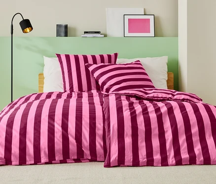 Lit avec des draps rayés rose et bordeaux, deux oreillers et un lampadaire.