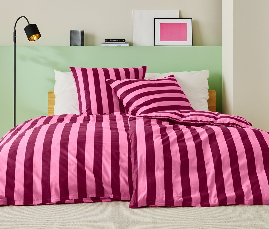 Lit avec des draps rayés rose et bordeaux, deux oreillers et un lampadaire.