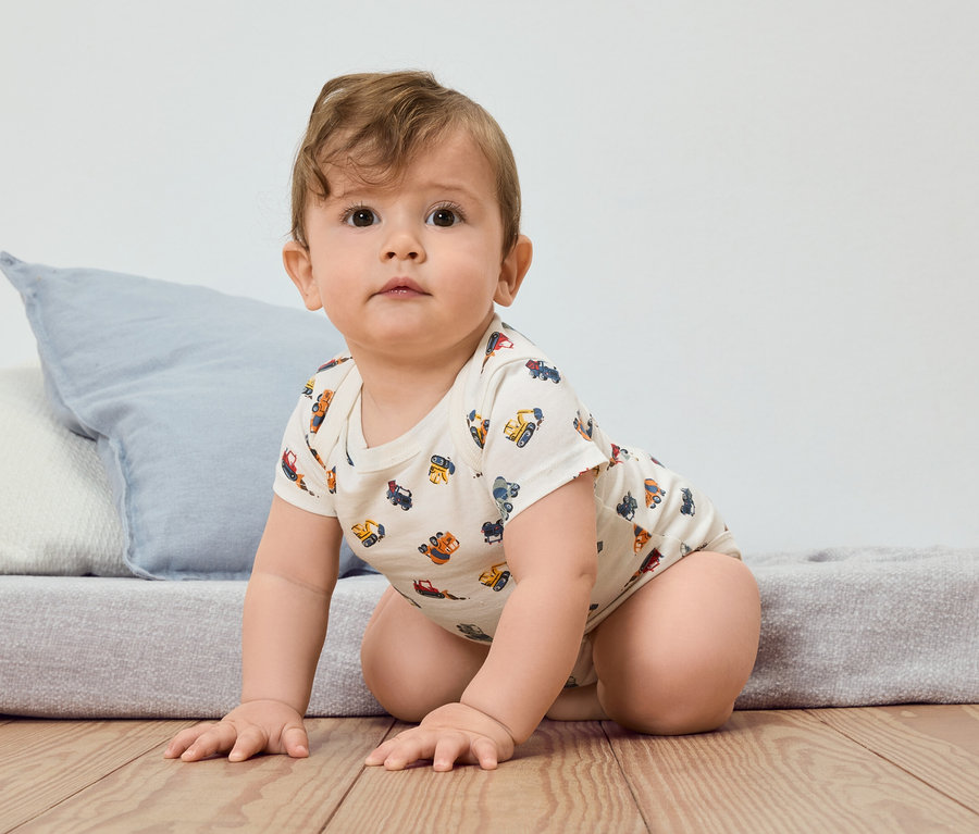 Un bébé rampe sur un plancher en bois, vêtu d'un body à manches courtes avec un motif de véhicules de construction.