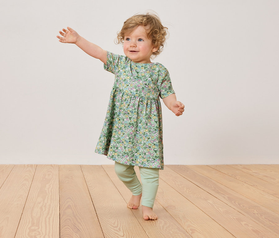 Un jeune enfant aux boucles blondes porte une robe à fleurs et des leggings verts, debout sur un plancher en bois.