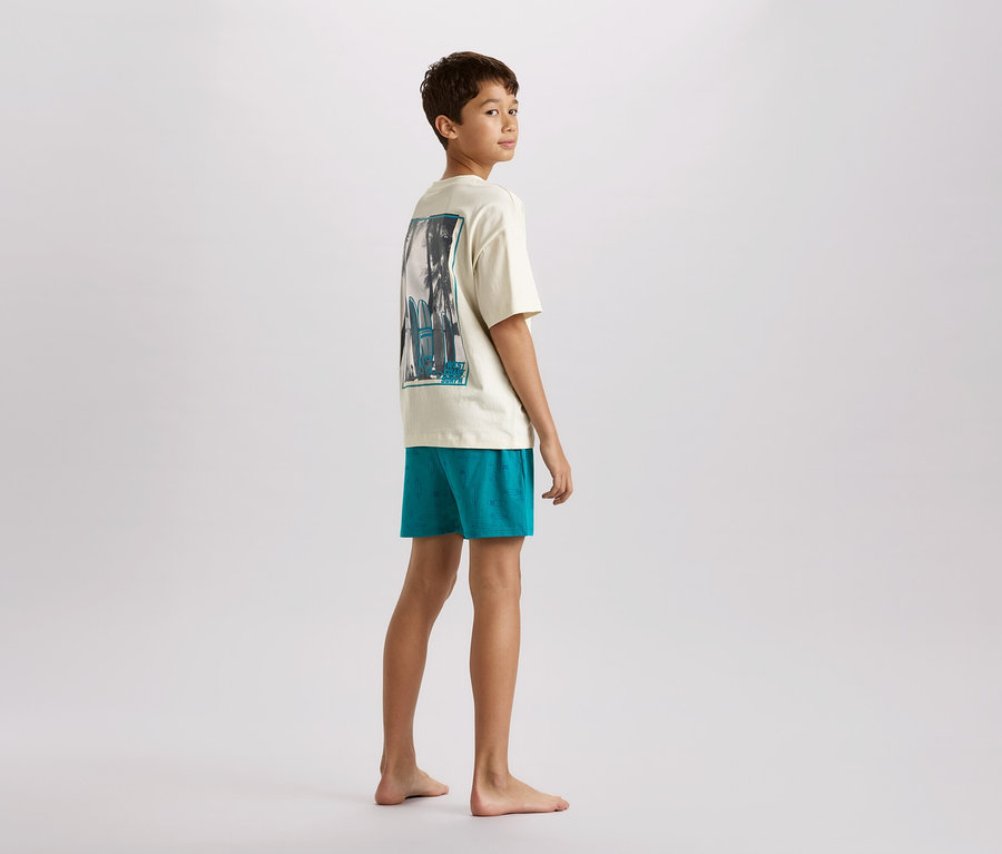 Un jeune adolescent se tient pieds nus et regarde par-dessus son épaule. Il porte un t-shirt crème avec un imprimé et un short turquoise.