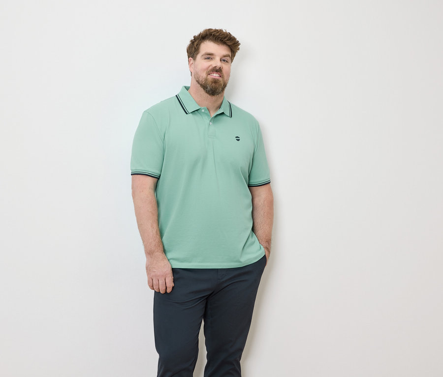 Un homme barbu portant un polo vert clair et un pantalon foncé devant un mur blanc.